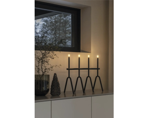 Dekorativt arrangemang med en svart lampa med fyra ljus, en liten gran och en dekorativ kvist i en interiör.