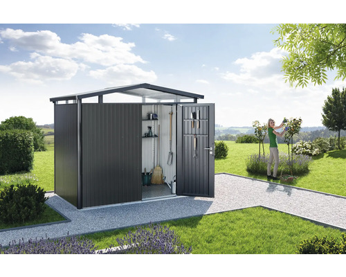 Trädgårdsskjul med öppen dörr i ett trädgårdslandskap med gräsmatta och en kvinna som beskär rosor.