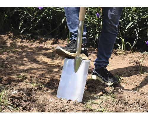 En person använder en spade med trähandtag i trädgården för att gräva jord.