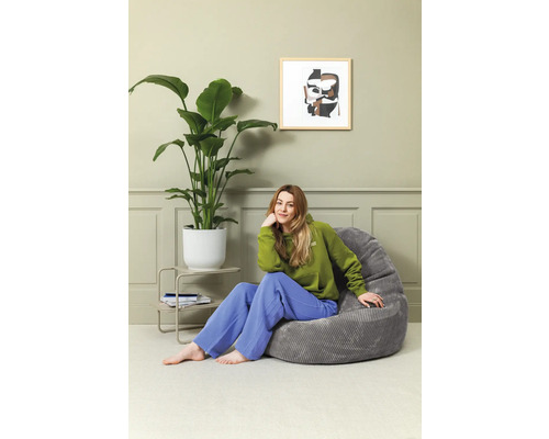 Kvinna sitter i ett rum på en grå sittsäck bredvid en krukväxt och ett sidobord