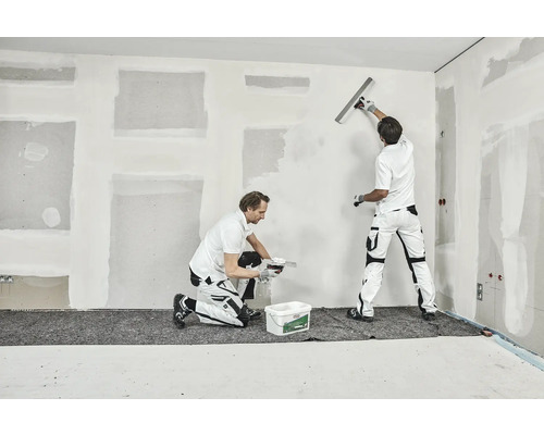 Två hantverkare spacklar en vägg i ett rum.