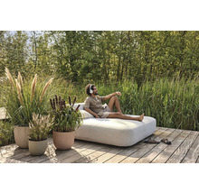 Utomhusscen med man som slappnar av med hörlurar på en solbädd i trädgården, omgiven av prydnadsgräs i planteringskärl på en träterrass.