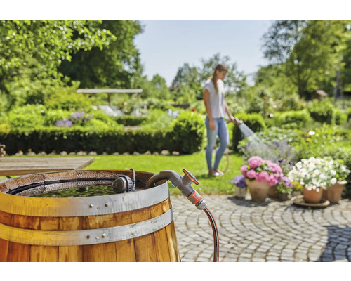 Trädgårdsscen med regntunna, kran och kvinna som vattnar blommor
