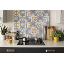 Organiserat ljust kök med ett stänkskydd med grå-gult kakelmönster ovanför en spishäll på en vit bänkskiva med tillbehör och växter.