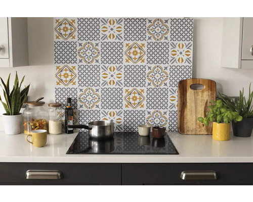 Organiserat ljust kök med ett stänkskydd med grå-gult kakelmönster ovanför en spishäll på en vit bänkskiva med tillbehör och växter.