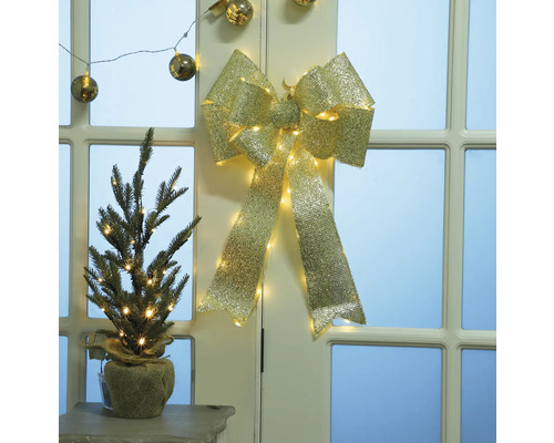 Dekorativ guldfärgad rosett med belysning och liten gran i jutesäck på bord framför dörr