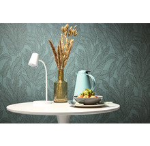 Dekorativt matbord med lampa, termoskanna, vas med torkade blommor och fruktskål framför en tapet med bladmönster