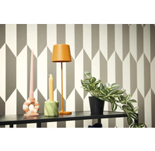 Dekorativ scen med lampa, ljus och krukväxt på en hylla framför en geometrisk tapet.