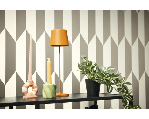 Dekorativ scen med lampa, ljus och krukväxt på en hylla framför en geometrisk tapet.
