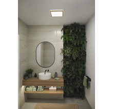 Ljust badrum med tvättställ, spegel, grön växtvägg och trähyllor för handdukar och badrumsartiklar under en fyrkantig taklampa.