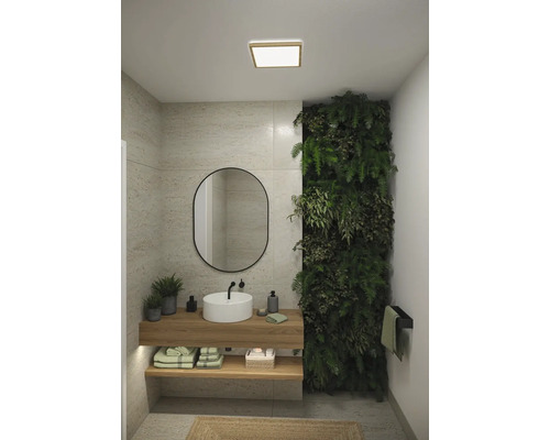 Ljust badrum med tvättställ, spegel, grön växtvägg och trähyllor för handdukar och badrumsartiklar under en fyrkantig taklampa.