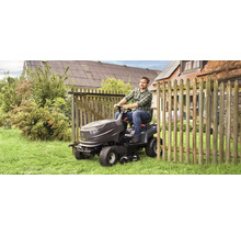 En man kör en trädgårdstraktor på en gräsmatta bredvid ett trästaket.