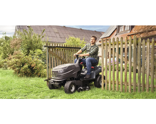 En man kör en trädgårdstraktor på en gräsmatta bredvid ett trästaket.