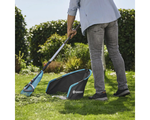 Trädgårdsscen med trädgårdsmästare som sopar gräsmattan med en lövräfsa och en gräsuppsamlare från Gardena