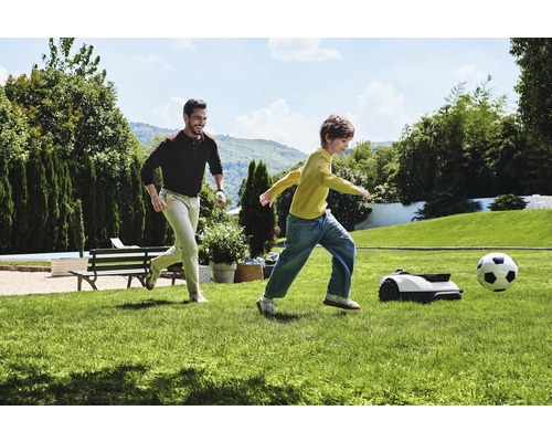 Man och barn spelar fotboll med robotgräsklippare i trädgården.