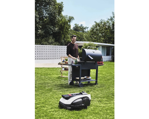Man grillar i trädgården med en grill och en robotgräsklippare.