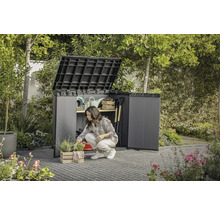 Trädgård med en kvinna som knäböjer framför ett öppet trädgårdsskåp där trädgårdsredskap förvaras.