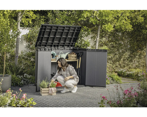Trädgård med en kvinna som knäböjer framför ett öppet trädgårdsskåp där trädgårdsredskap förvaras.