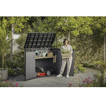 Trädgårdsscen med redskapsbod, trädgårdsredskap och en kvinna som håller en låda med växter.