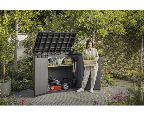 Trädgårdsscen med redskapsbod, trädgårdsredskap och en kvinna som håller en låda med växter.