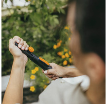 En person håller en trädgårdsslang med sprutmunstycke mot en bakgrund av gröna växter och gula blommor.