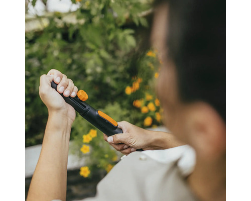 En person håller en trädgårdsslang med sprutmunstycke mot en bakgrund av gröna växter och gula blommor.