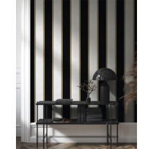 Rumsöversikt med randig tapet i svart, vitt och guld samt hylla med böcker och lampa.