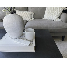 Dekorativt arrangemang på ett bord med vas, ljus, bok och fjärrkontroll framför en soffa.