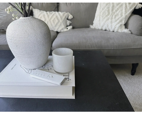 Dekorativt arrangemang på ett bord med vas, ljus, bok och fjärrkontroll framför en soffa.