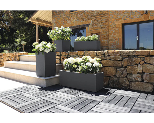 Utomhusbild av rektangulära och kvadratiska planteringslådor med blommor framför en byggnad.