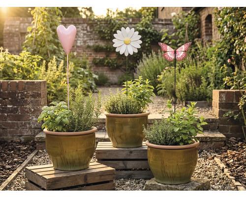 Tre blomkrukor med örter och dekorativa trädgårdspluggar i form av ett hjärta, en blomma och en fjäril i en trädgård.