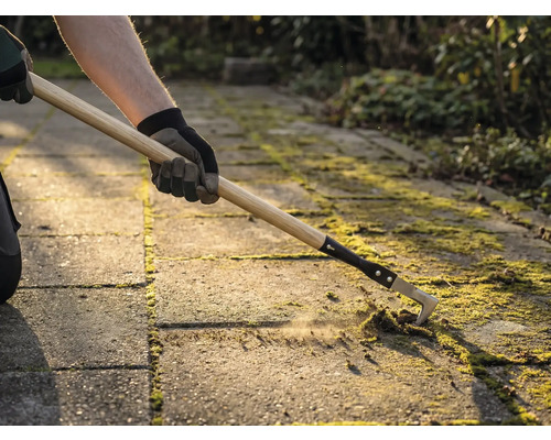 En person tar bort mossa mellan stenplattor med en fogkrats.