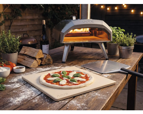 Utomhusbild av en Ooni pizzaugn med pizza, pizzaspade och ingredienser på ett träbord.