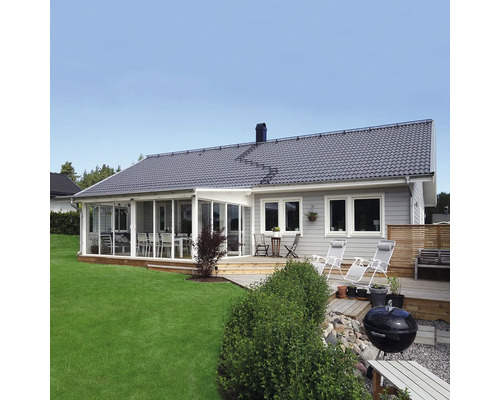 Trähus med grå fasad, stort inglasat uterum och ett trädäck med solstolar och grill på en gräsmatta.