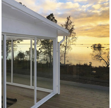 Vitt uterum med stora glasskjutdörrar på en trätrall med glasräcke och utsikt över en sjö vid solnedgång.