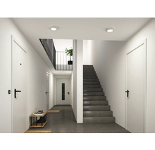 Modern ljus hall med mörk trappa, vita dörrar med svarta handtag och runda takspotlights.