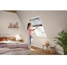 En kvinna justerar en mörk plisségardin på ett vitt takfönster i ett ljust och modernt sovrum med snedtak.