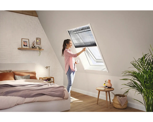 En kvinna justerar en mörk plisségardin på ett vitt takfönster i ett ljust och modernt sovrum med snedtak.