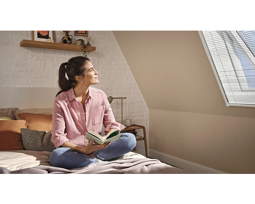 Kvinna läser på en säng i ett ljust vindsrum med takfönsterpersienn, vit tegelvägg och hylla i trä.