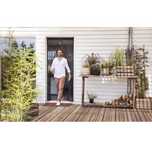 En man går genom ett magnetiskt insektsnät vid en altandörr ut på en träterrass med många krukväxter.