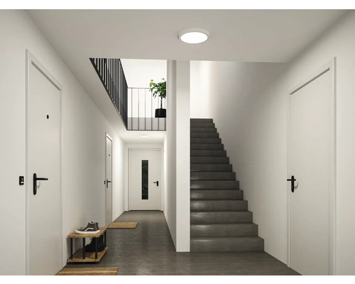 Modern, ljus hall med rund taklampa, grå trappa, vita dörrar och svart räcke.