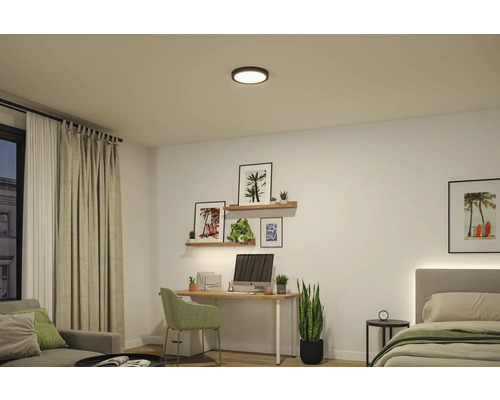 Modern, ljus inredning med en rund svart LED-taklampa över en arbetsplats med skrivbord, stol och en säng.