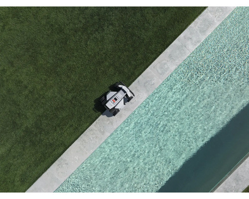 Robotgräsklippare vid kanten av en gräsmatta bredvid en pool med stenram, vy uppifrån.