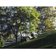 Robotgräsklippare med fyrhjulsdrift klipper en brant grässlänt under stora gröna träd i en trädgård.