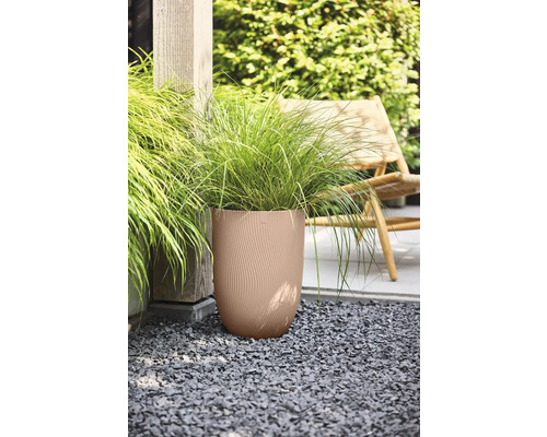 Hög rund planteringskärl med vågig struktur och prydnadsgräs på grus i en trädgård bredvid en trästolpe och en trädgårdsstol.