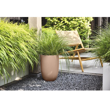 Brun rund blomkruka med räfflad struktur och grönt prydnadsgräs på en terrass med mörkt grus bredvid en trästol.