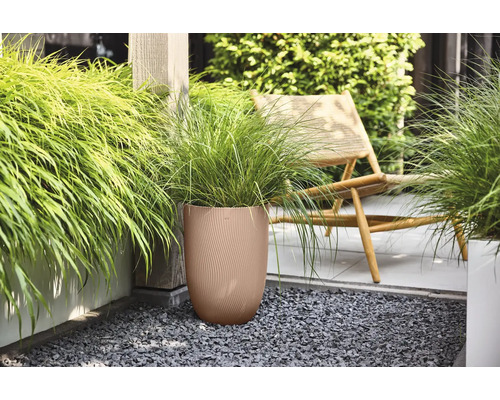 Brun rund blomkruka med räfflad struktur och grönt prydnadsgräs på en terrass med mörkt grus bredvid en trästol.