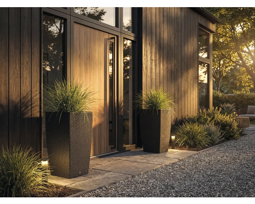 Två höga, mörka planteringskärl i rottinglook med prydnadsgräs vid en modern, organiserad entré med träpanel och stengång.