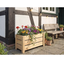 Rektangulär blomlåda i ljust trä fylld med färgglada blommor, framför en korsvirkesvägg på en stenterrass bredvid en träbänk.