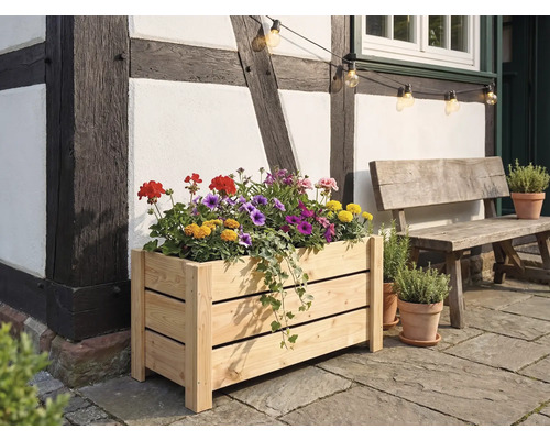 Rektangulär blomlåda i ljust trä fylld med färgglada blommor, framför en korsvirkesvägg på en stenterrass bredvid en träbänk.